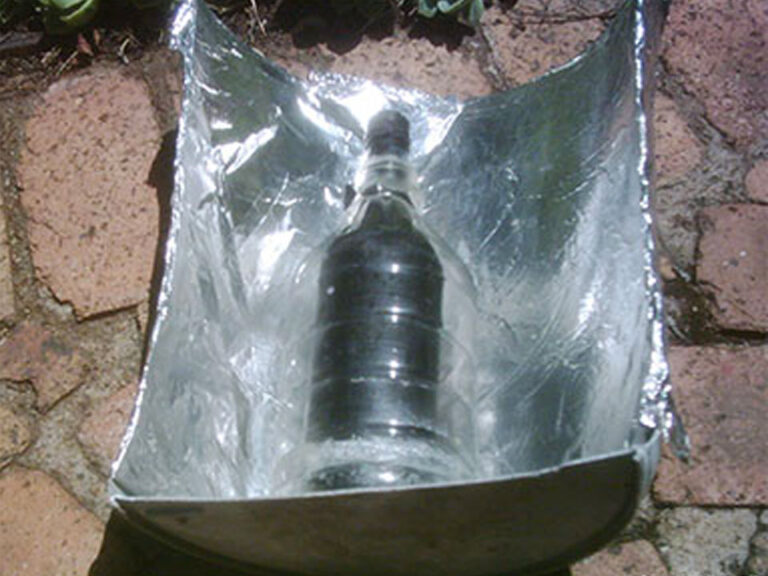 A bucket cut in half lengthwise, lays on its side. The inside of the bucket is lined with aluminum foil.  Inside the bucket is a clear plastic bottle. Inside this bottle is a capped dark black bottle.