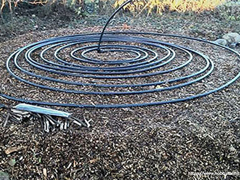 A brown dirt or ground compost patch with a spiral of black tubing on top. In the middle of the spiral the tube rases up into the air.