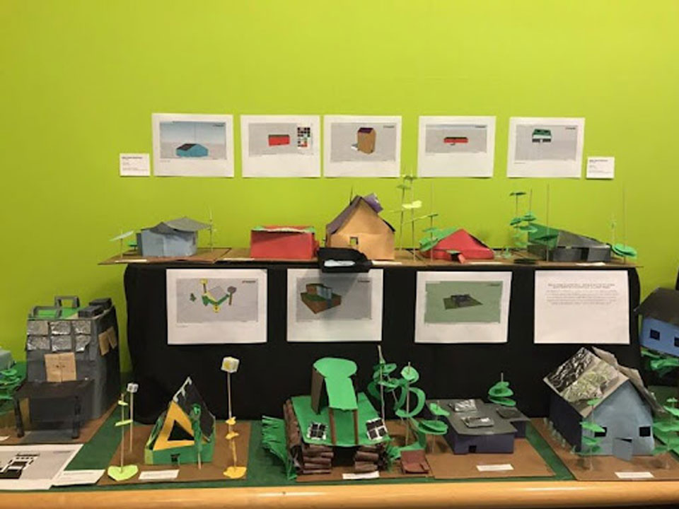 A green wall with five printouts of building models. Each building is made of paper and on a black table underneath it's corresponding image. On a dark green topped table with wooden border under the black table are four more papers and models. On the left, still on the green table, is another model with the paper in front of it. Each structure is different and made from different colored paper.