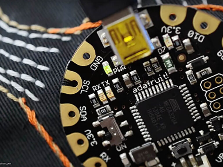 Adafruit FLORA  board on a black fabric. on the left side 5 lines of grey running stitches are seen between two lines of orange and brown backstitched lines.