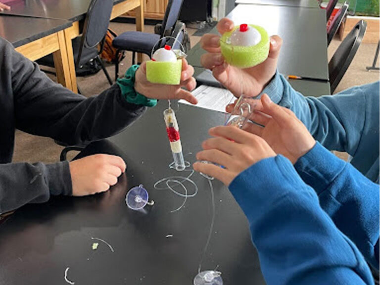 Student holds up lime green foam circles attached to clear plastic tubes wrapped in red wire in the middle between two strips of masking tape. The green foam have a fishing bobber in the middle. A suction cup with fishing line can be seen on the table.