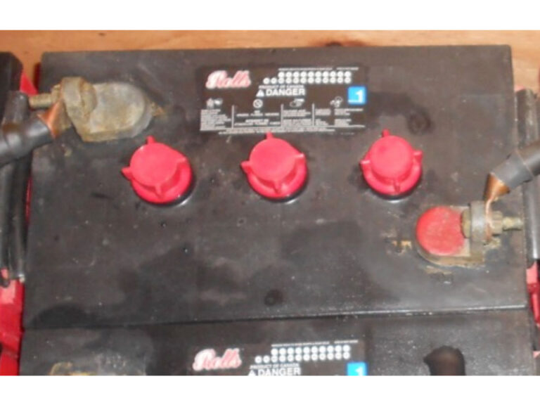 A close image of the top of a car battery. On the upper left is the negative terminal connection with a black cable in the middle are three red cell access caps and in the bottom right the positive terminal with a black cable connected.