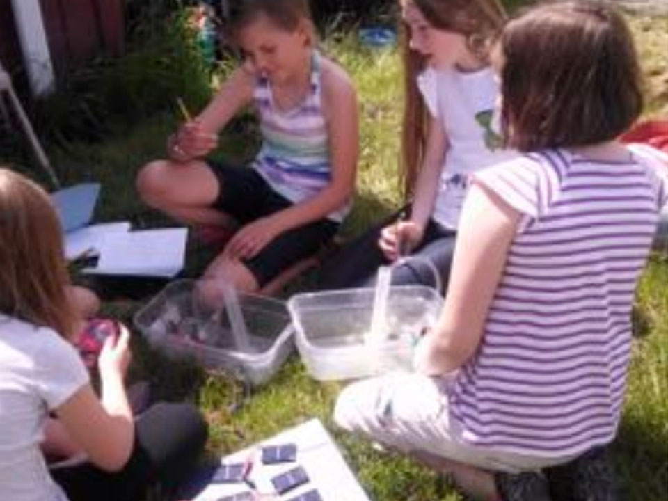 4 girls sit in a circle on grass with two plastic tubs. Each tub has a graduated cylinder one has a clear plastic tube coming from the top. Next to the girls is a paper with small solar panels connected together.