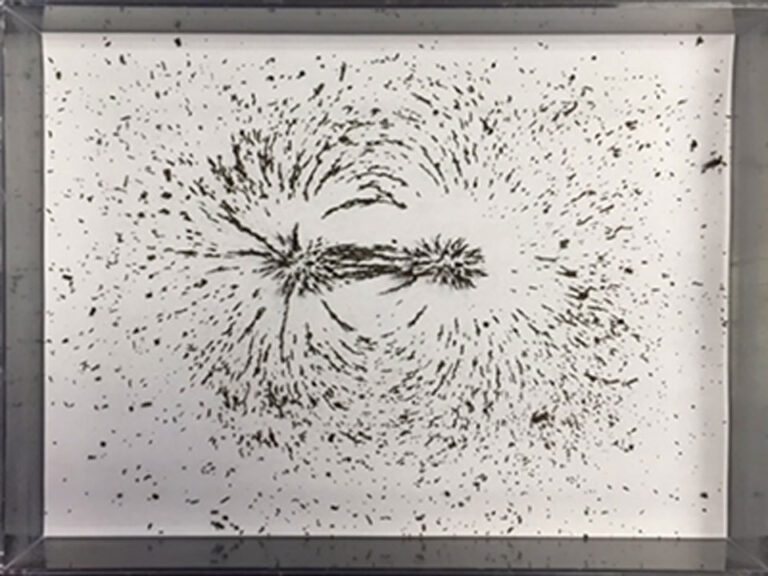 Picture of a black framed shadow box with white paper inside the frame.  On the paper are metal shavings that are dispersed in two circular patterns reflecting the electromagnetic field being generated by an unseen device.