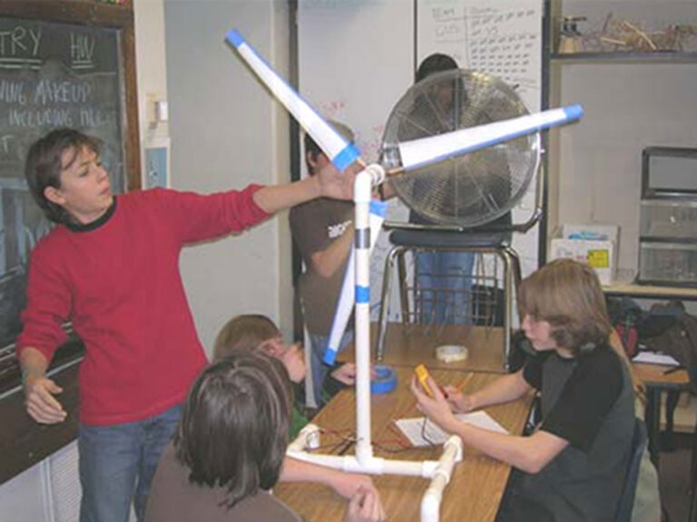 Photo of 6 students us a a metal fan to test a handmade wind turbine sitting on top of a desk. The base and shaft of the turbine are made of PVC. It has three white triangular blades with blue tape on the ends and one side holding them to dowels.