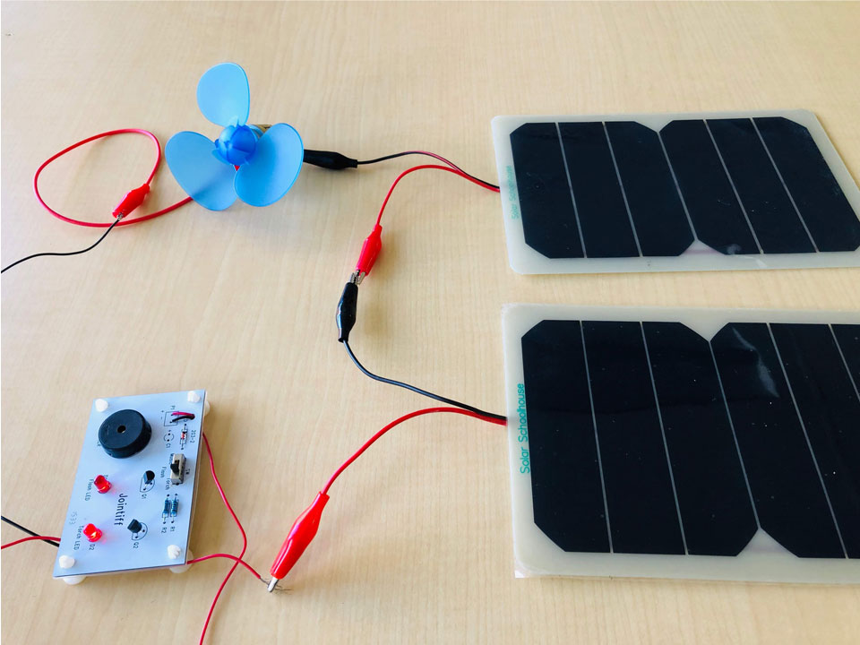 On the right two solar panels are connected together using an alligator clip. The lower panel's red wire connects to a sound and light board with two LEDs, resisters, a switch, and a speaker, The connecting wires from the board go to the top left of the images where a blue fan is connected. The circuit closes with the top solar panel.