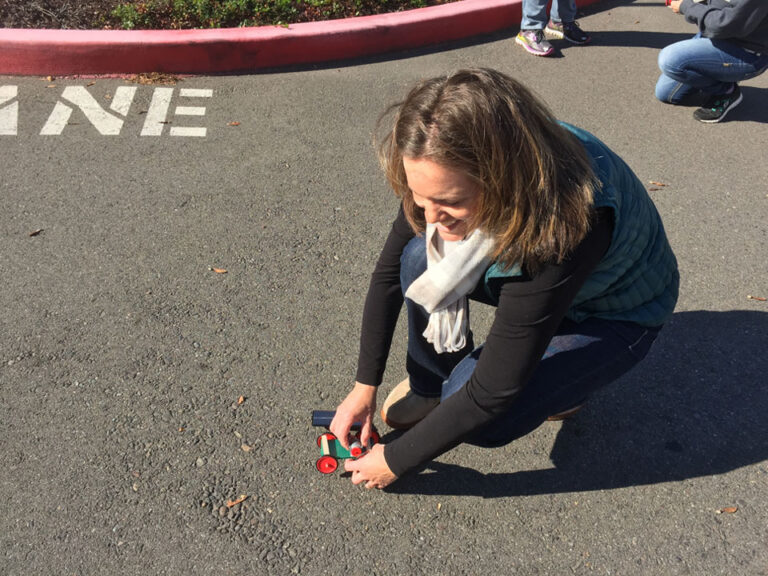 On the blacktop on a sunny day the picture looks down onto a kneeling woman setting a model solar car onto the pavement.