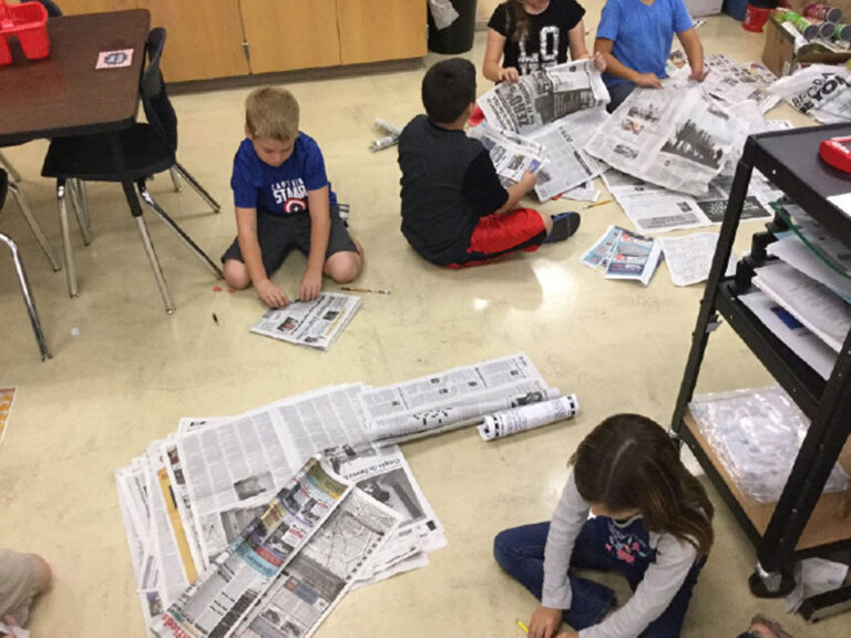 Students sit on a linoleum floor with piles of newspapers which they are folding and rolling with pencils.