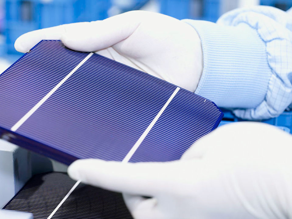 White gloved hands hold a dark blue solar panel with two silver bands across it. Below is another solar panel laying down on an unseen surface.