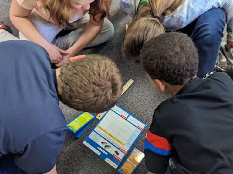 Students sitting in a circle on grey carpet with a printout, a ruler, and pencils.