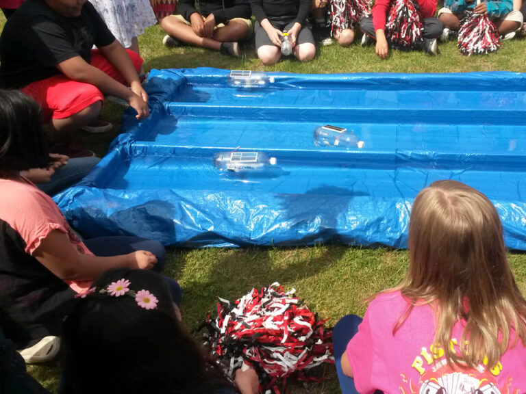 A shallow rectangular pool is divided into three lanes by two raised dividers and lined with a tarp. In each lane a solar boat made from a clear plastic water bottle can be seen.