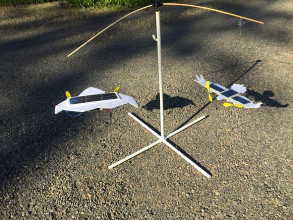 A white mobile with two arms. Hanging from the left arm is a solar powered paper plane. The solar panel is on the top and 2 propellors. On the right arm is a bird shaped paper plane with 3 solar panels down wing and on the back and 2 propellors.