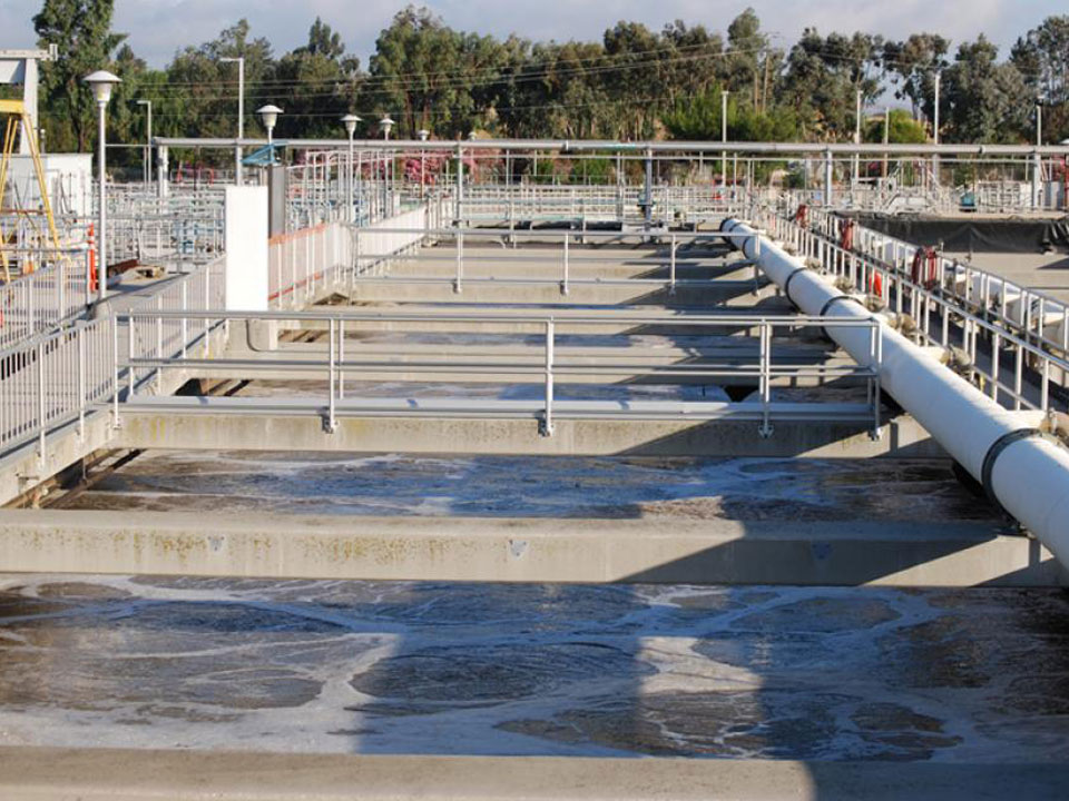 Picture of a wastewater tank at a wastewater treatment facility. The water is frothy and dark. A white pipe runs from the top center to the bottom right of the photo over the tank over the top of concrete supports.