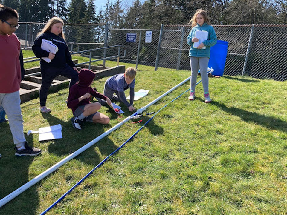 A photo of a green field. From the top center to the bottom left is a a water pipe. At the top of the photo the pipe is connected to a water storage tank. Students kneel on the left side of the pipe.