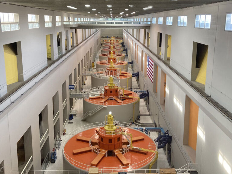 A large room with a row of hydro turbines with orange caps running it's length. Pipes connect each turbine to the wall on the right.