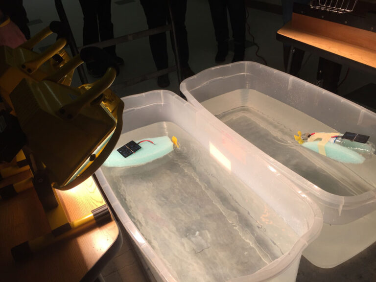 Two bins of water each with a solar boat model, under halogen lights. One boat is made of a plastic bottle with two oval green foam pieces, a small solar module, and a propellor in the opening. The other is a green foam boat with a propellor and a solar panel.