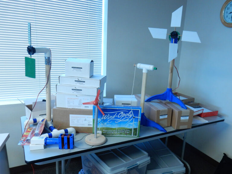 A table covered in boxes and 4 handmade wind turbine models; one has 2 rectangular green blades, one has 4 white blades, one has no blades, and the smallest has a red 3 blade propeller. Behind the red turbine is a blue box with white writing that reads "Wind-opoly"