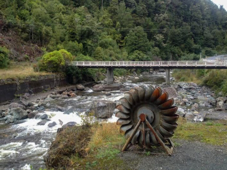 On the bank of a river is a metal water turbine with rust on many of the blades and on the A-frame stand. Nearby the river runs with shallow rapids and a bridge in the background.