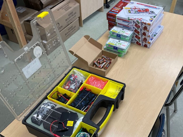 A table with an electronic kit with the lid open. It has small solar modules and wires. Next to it are; a cardboard box with AA batteries, a plastic kit with a white lid and green contents, and a stack of white and red boxes of Snap Circuits.