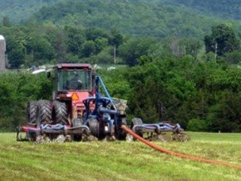 Photo shows a red tractor with a 7-blade plow attachment working through a green field. In the middle of the attachment a red hose lays out in the field.