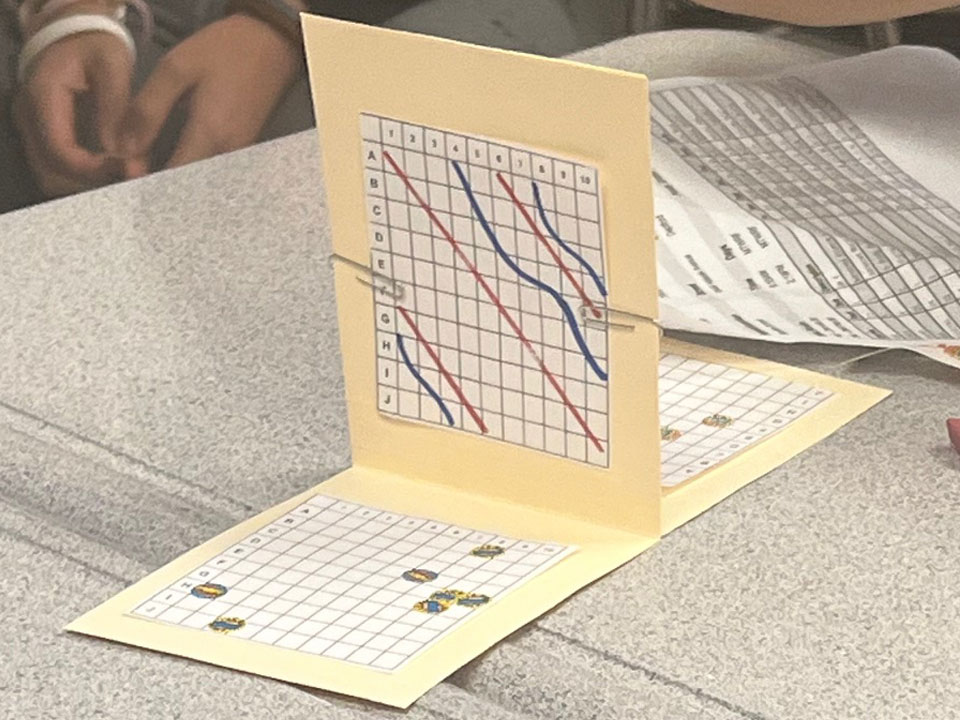 T-shaped piece of paper with a grid on the table surface in the foreground and a grid with diagonal red lines and curvy diagonal blue lines on the vertical surface. The back surface also has a grid. The game sits on a grey classroom desktop.