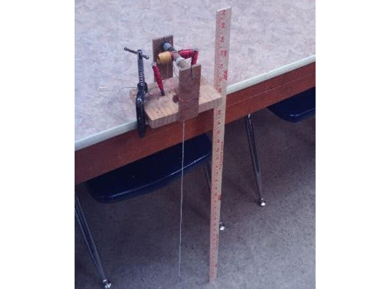 A handmade electric motor clamped to a table. The motor is a dowel has two horizontal wings that are wrapped in copper wire that are resting parallel wooden base. Nailed into the wood on each side is a nail wrapped in copper wire. A string is connected to the dowel with a weight to make the dowel spin. A yardstick leans next to the table the measure is unreadable, but most of the stick.