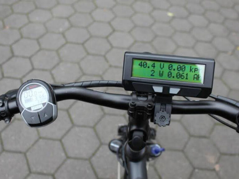 Photo of bicycle handlebars. On the left side of the handlebar is mounted stopwatch device. Just right of the middle of the bars is a screen which reads 40.4 V 0.00kph on the top line and 2 W 0.061Ah on the bottom line.