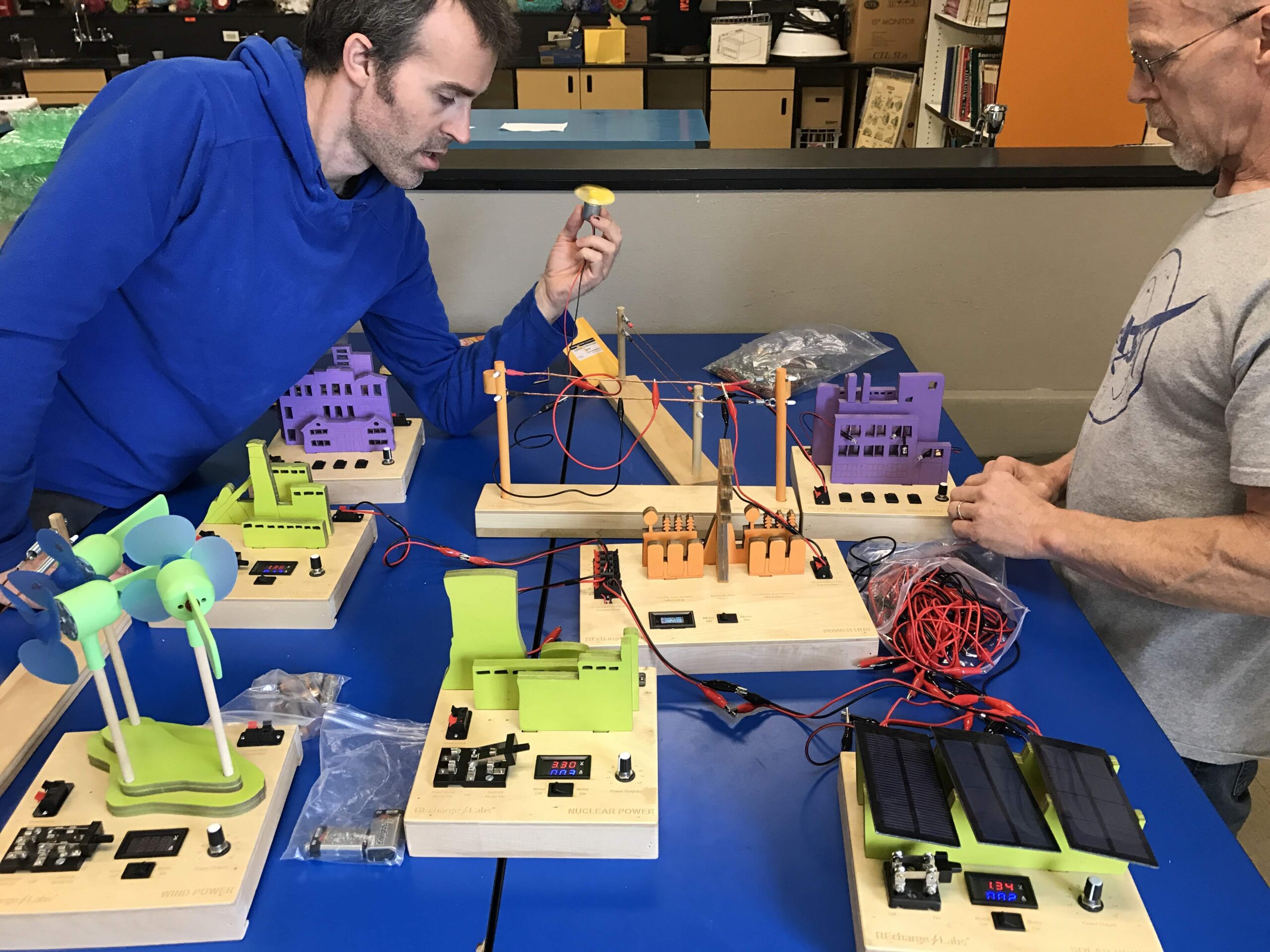 2 men sit at a blue table with a power grid model. The man on the left holds a motor with yellow fan that is spinning. It is connected by alligator clips to a model power line. From front to back there are models of wind power, with three KidWind fireflies, a nuclear reactor, a solar farm with 3 solar modules, a transfer station, a coal plant, a residential area, and an industrial area.