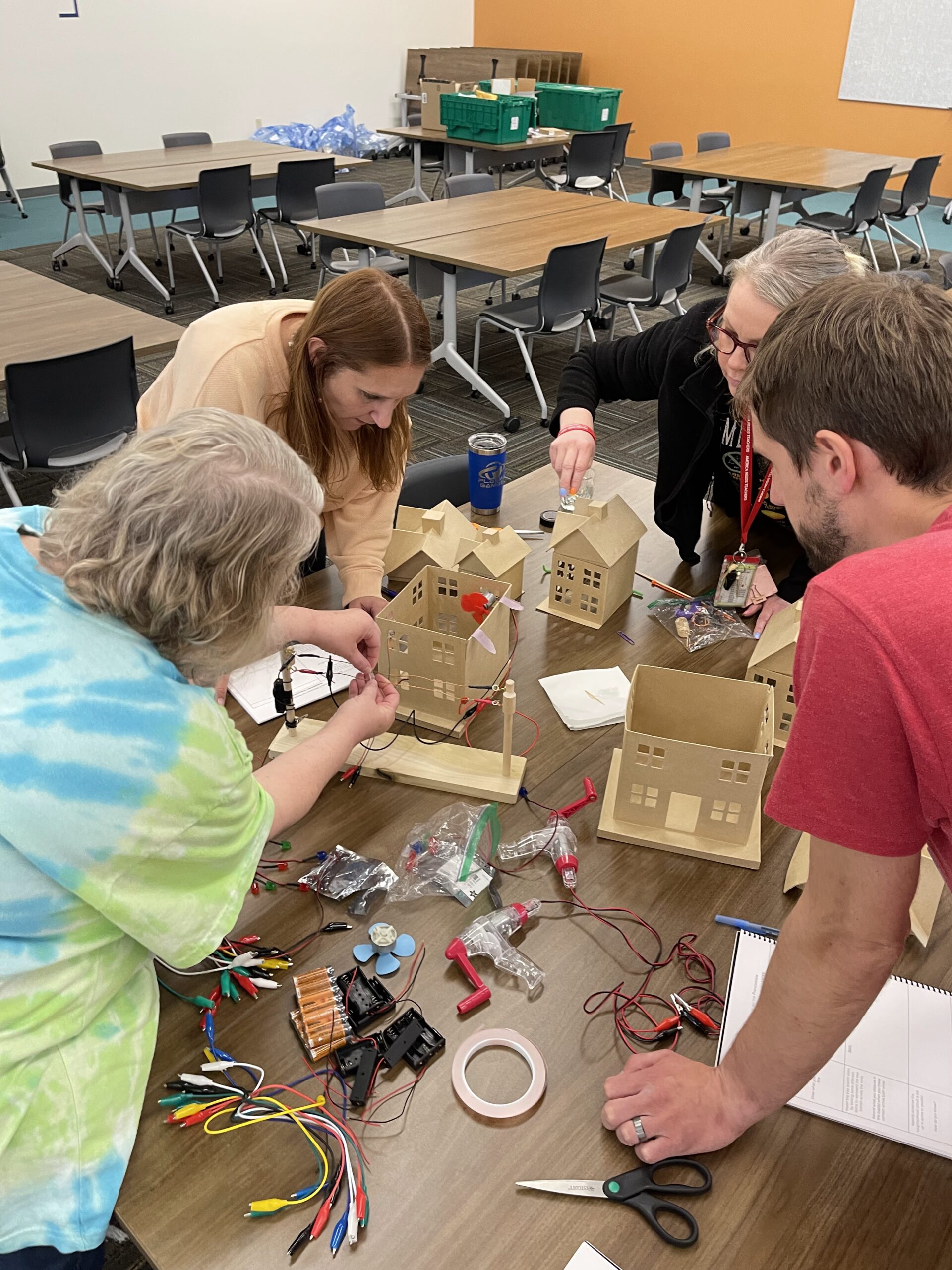 4 adults work around a table connecting alligator clips to a model power line to power lights and fans they have connected to paperboard model houses.