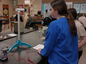 Two students stand at a lab table with a Kidwind wind turbine model connected to a multimeter.