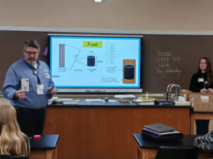 Teacher shows a card to the class while a screen shows fuel pellets, how they are assembled into rods and fundamentals of fusion