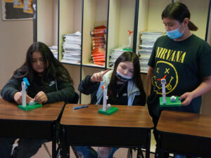 3 students at desks have wind turbine models with a green base, white tower with a motor at the top and a propellor (either red or blue). On the green tray is a box that the wires from the motor connect to.