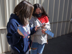 2 girls stand in the sun each holding a solar module connected to a handheld device they are looking at.