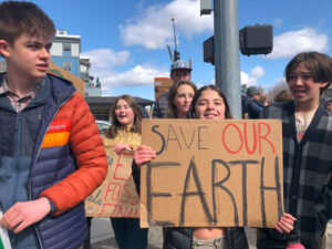 Students at a crosswalk hold a cardboard sign that reads, "SAVE OUR EARTH".