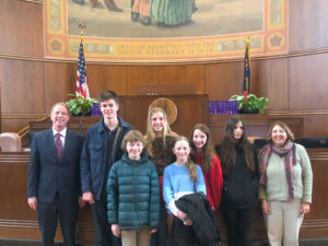 A legislator a group of students and a teacher stand in front of the bench at the Oregon Legislature posing for the group photo