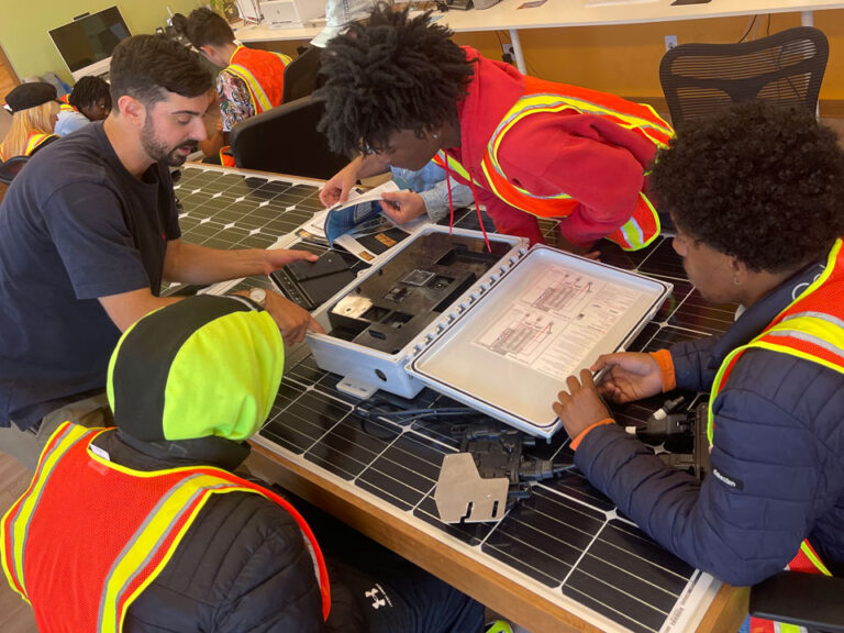 An educator and three young people in visibility vests lean over solar panels on a table, on top of which is a white case and several booklets.