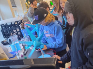 A group of students stand at a table with two blue wind turbine models. One student is using a multimeter.