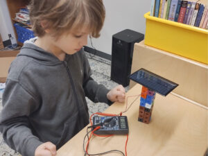 A young boy is using a solar module elevated on math blocks and connected to a multimeter which reads 0.56