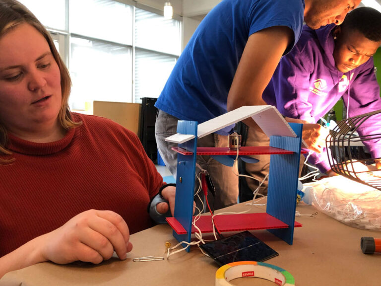 Teacher uses a model structure made of corrugated plastic. The model has a solar panel connected with alligator clips to a motor and fan in the model ceiling.