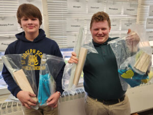 Two boys hold wood, pipes, dowels, and plastic pieces to assemble Kidwind turbine models.