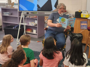 Teacher sits in a chair and reads from Everyday Superheros to a group of seated girls.