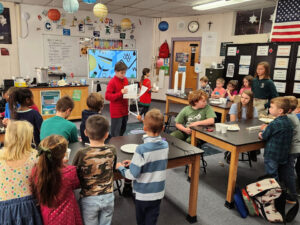 Student holds a paper in front of a class of mixed age students who stand and sit around lab tables.