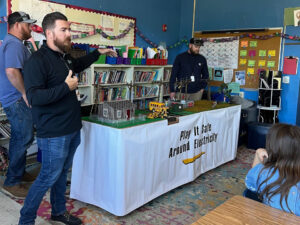 Three bearded men present a table with a model house, construction equipment, school bus, field, and powerlines running the length. The white table cloth says Play It Safe Around Electricity. A