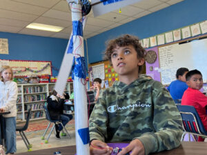 A student looks up at a wind turbine while connecting a 9V battery at the base. Other students in the room look at the turbine.