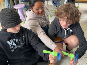 Three students hold 2 Kidwind fireflies on top of pencils. their blue propellors are spinning.