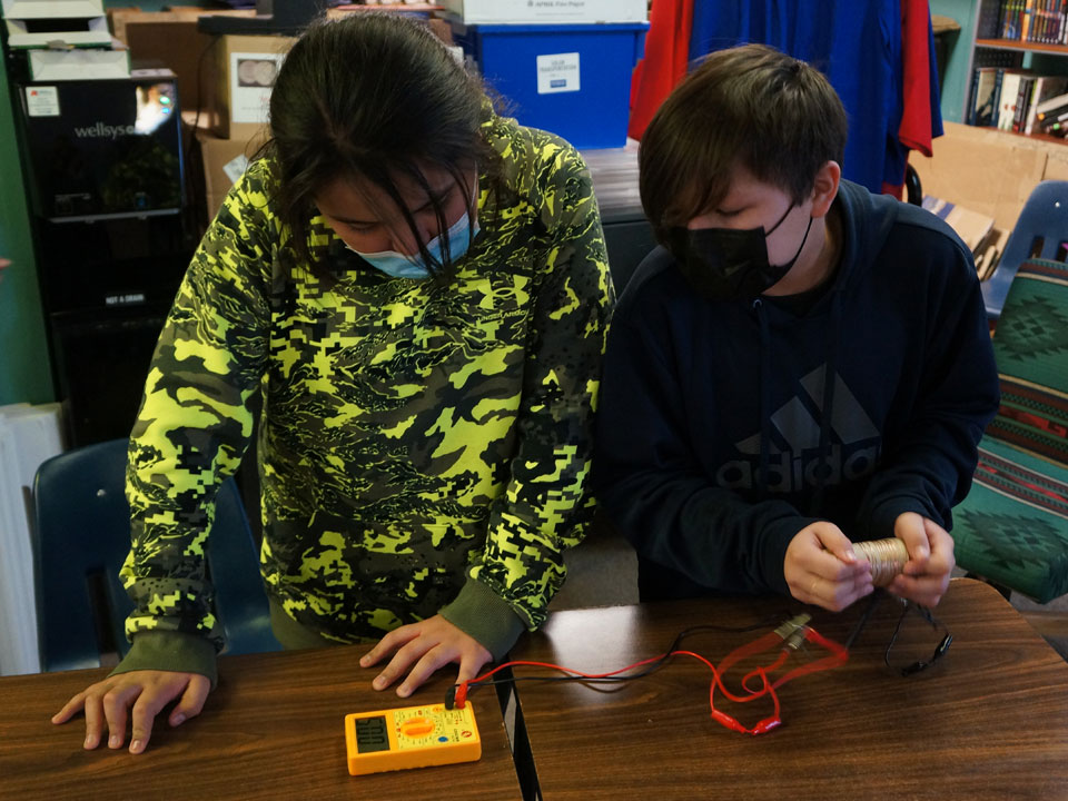 Two students use a multimeter to measure energy created by a wire wrapped cardboard tube being moved by the student on the right.