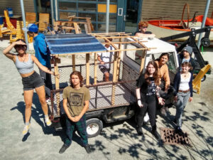 Students stand around a small truck with a solar panel on the top of the back cargo compartment.