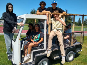 Students sit in a small truck with a wooden frame over the cargo area. It is parked on a green field.