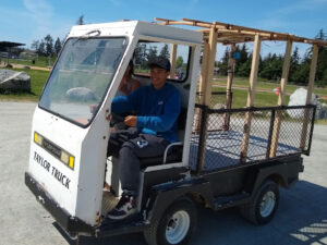 2 students drive a small truck with the words "TAYLOR TRUCK" on the front the cargo area has a wooden framework over it.