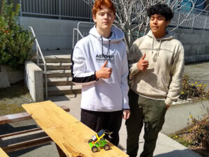 2 boys stand in the sun giving a thumbs up. On a bench in front of them is a small solar car model with a solar module and 2 AA batteries.